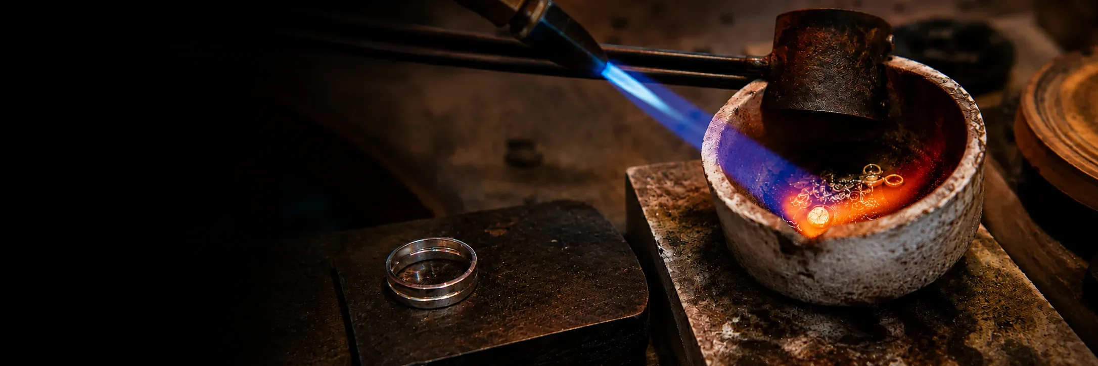 Gold melting in a crucible under a jeweller's torch, the metal glowing orange and the flame blue, with a finished band resting on a steel block alongside.