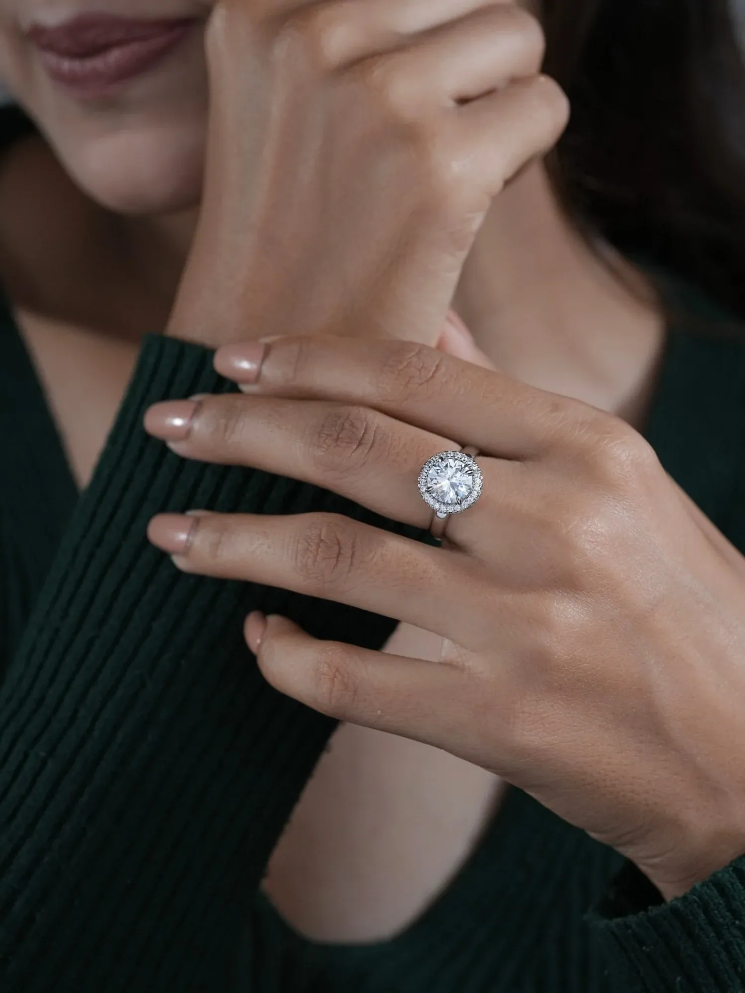 A hand wearing a round halo ring, held against a dark green ribbed jumper.