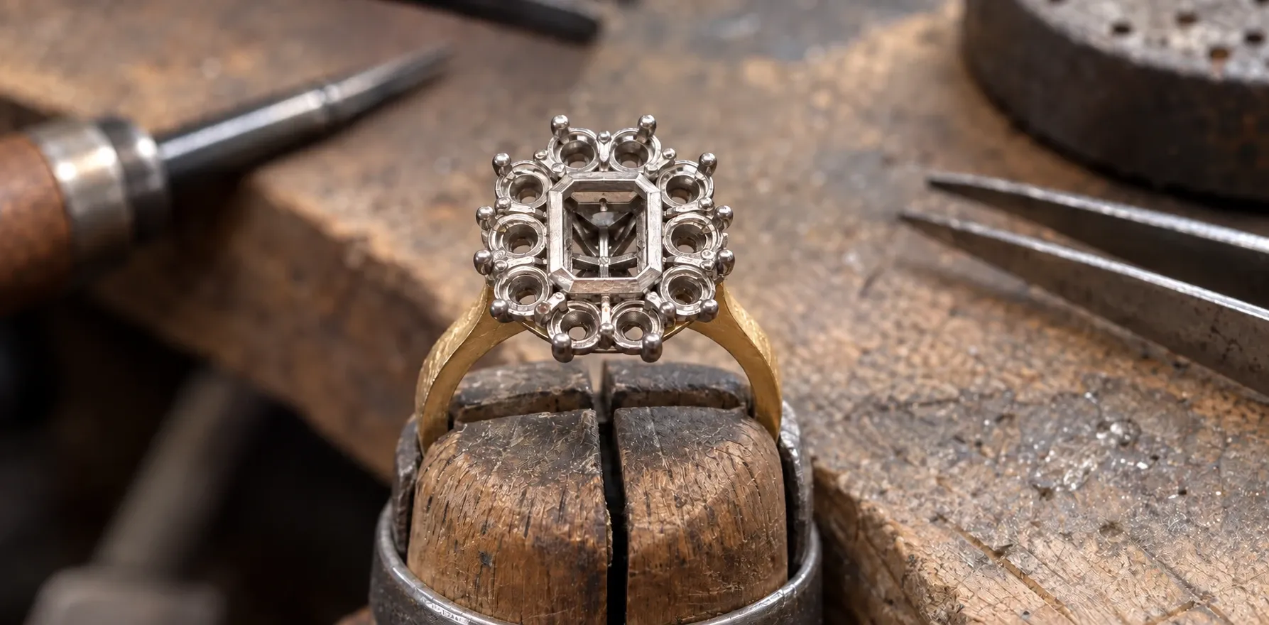 The cluster ring's empty mount in a ring clamp: an emerald-cut centre seat inside ten round seats, every claw standing, on a yellow gold band not yet polished.